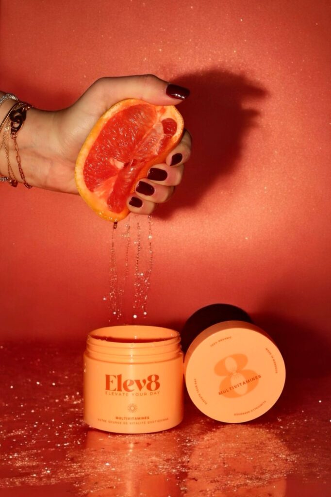 Hand squeezing grapefruit juice above Elev8 multivitamin jar on red background – representing freshness and natural vitamin supplementation.
