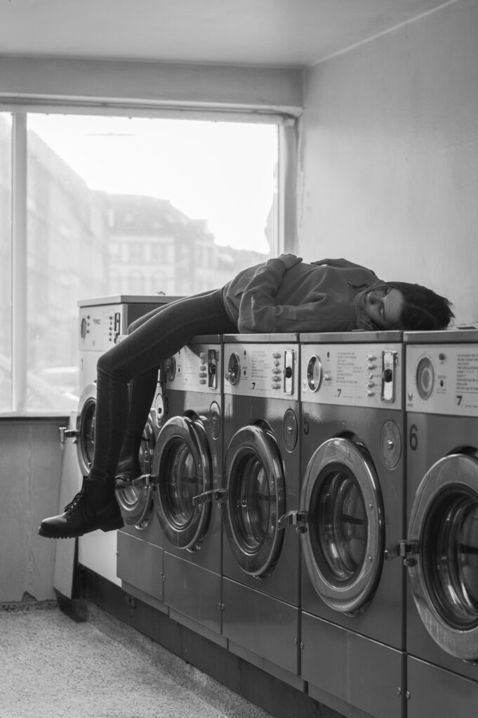 Exhausted woman sleeping on washing machines in laundromat – chronic fatigue and burnout representation